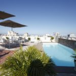 Piscina com barraca de sol. Arbustos e vista para outros prédios