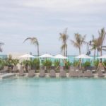 Piscina com cadeiras de esteira, barracas de sol e coqueiros atrás